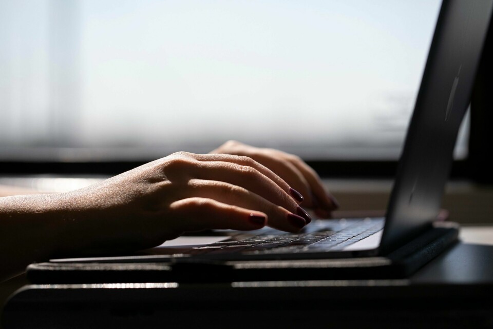 Illustrasjonsfoto. To hender med mørk neglelakk skriver på tastaturet til en laptop. Laptoppen vises fra siden, og hendene er glatte. Det er et vindu bak og lyset gir en litt selsom fornemmelse.