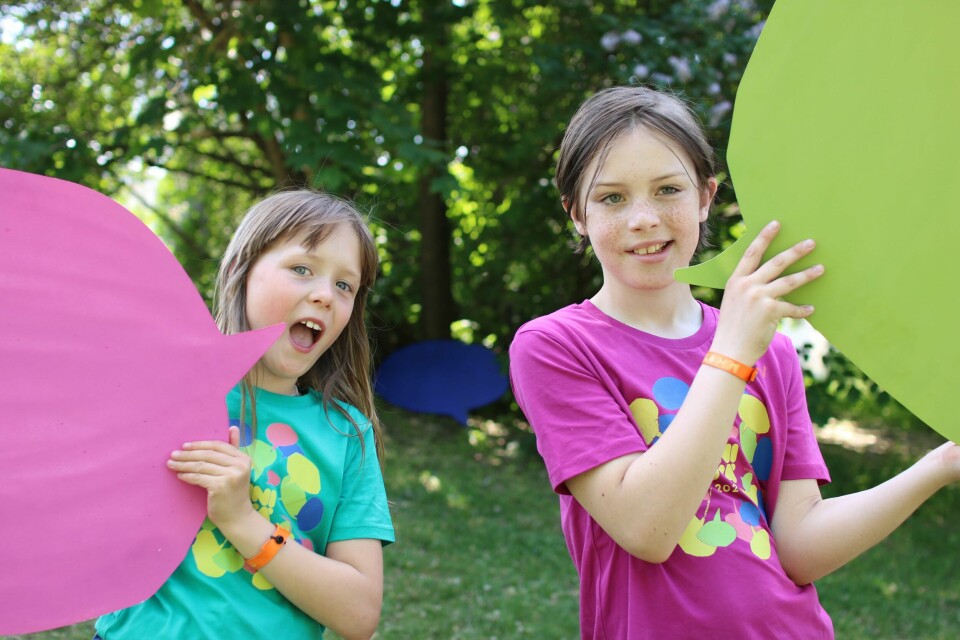 Eva Lunder Carlhovd (9) og Alfred Riiser Olsen (11) er sjefer for Miniøya 2023. To barn som har på fargerike t-skjorter og holder to snakkebobler i hendene.