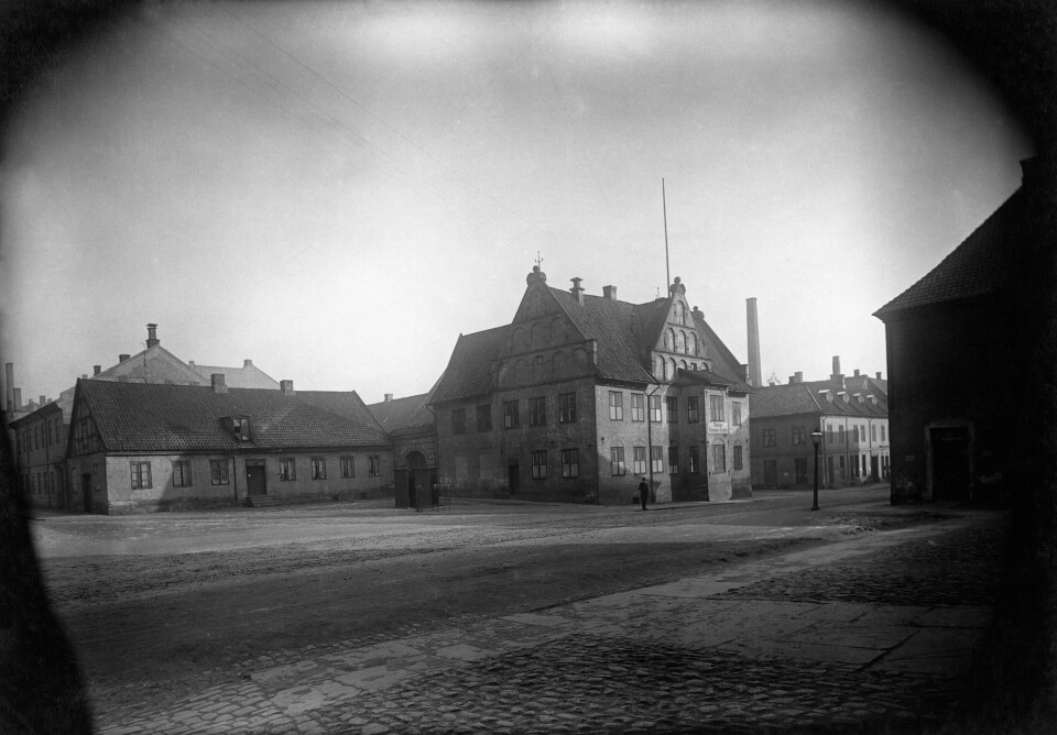 Christiania torv med byens eldste gjenværende hus i kvadraturen til høyre, Rådhusgata 19, kalt Garnisonssykehuset eller Rådmannsgården, fra 1626. Her bodde borgermester og kjøpmann Niels Lauritzen og kona Margrethe Lauritzdatter da Anna Rhodius anklaget henne for å være en «forhenværende hore» og borgermesteren for å skaffe seg inntekter på ulovlig vis. Foto fra 1896. Folketom brosteinbelagt plass med bygninger i bakgrunnen.