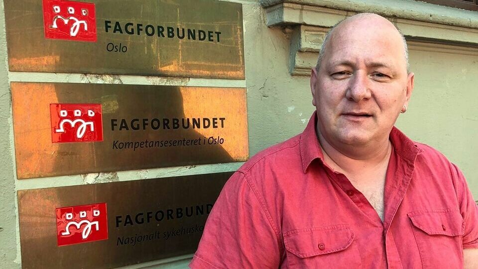 Roger Dehlin er leder for Fagforbundet Oslo. Skallet mann i rød skjorte står foran en murvegg med gullskilt på seg. Det står fagorbundet på de.