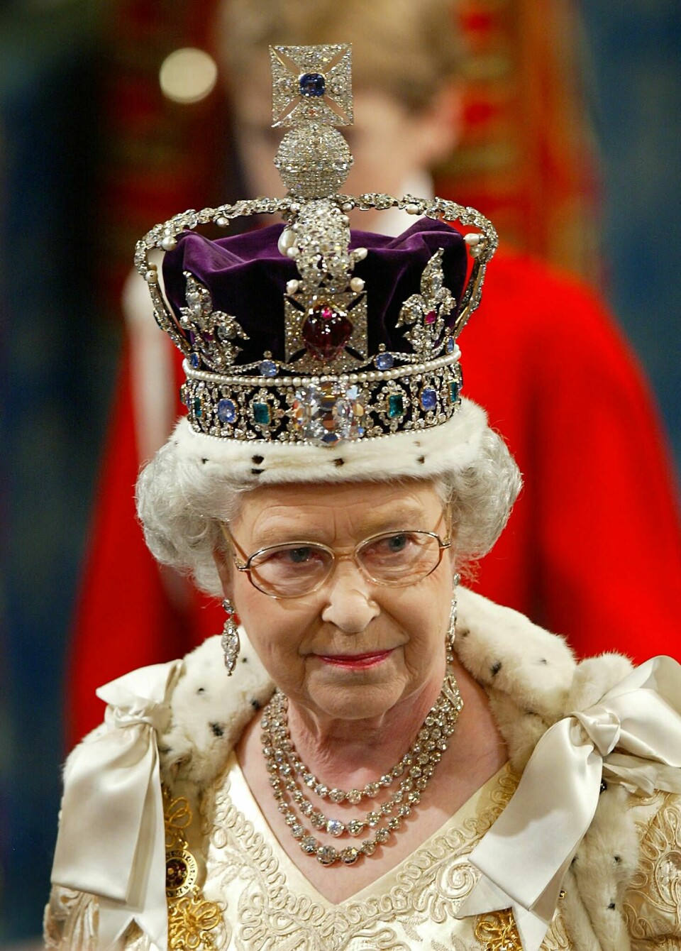 Dronning Elizabeth II av Storbritannia iført dronningmorens krone. Dronning Elizabeth i godt voksen alder. Hun har på briller, hvit kjole, kappe og en krone i lilla. Kronen har en pelskant, juveler og en greie som stikker opp.