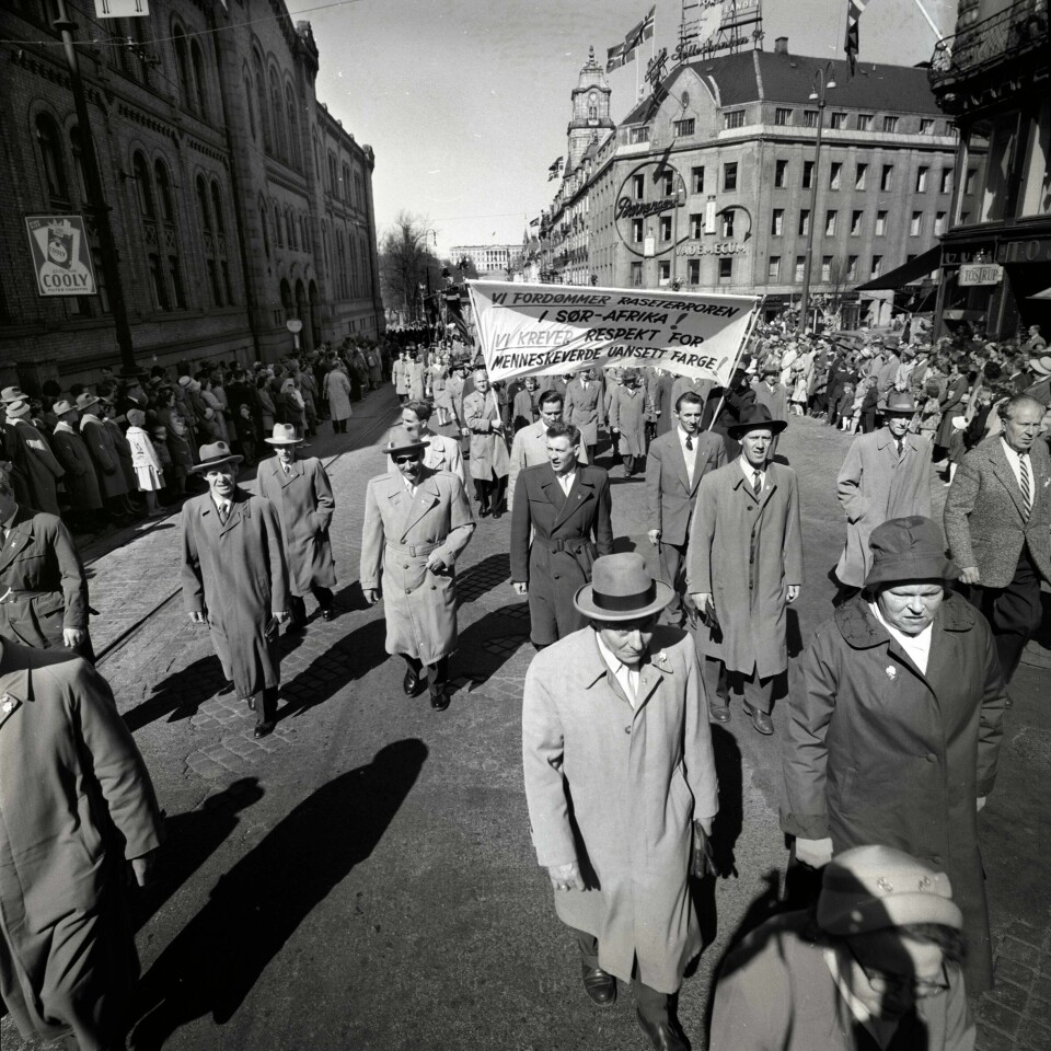 1. mai-toget i 1960 på Karl Johans gate med parolen «Vi fordømmer raseterroren i Sør-Afrika! Vi krever respekt for menneskeverdet uansett farge!» Mange folk står på fortauet og ser på demonstrantene som går opp langs Karl Johans gate.