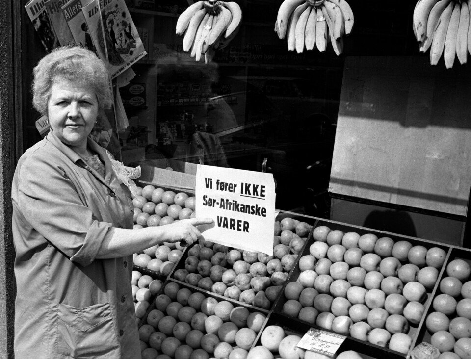 I April 1963 innførte Sør-Afrika nye drastiske apartheid-lover. Folk demonstrerte over hele verden mot apartheid-regimet og krevde boikott. Denne Oslo-butikken har juni 1963 skilt i vinduet «Vi fører IKKE Sør-Afrikanske varer». En dame peker på et skilt i et butikkvindu.