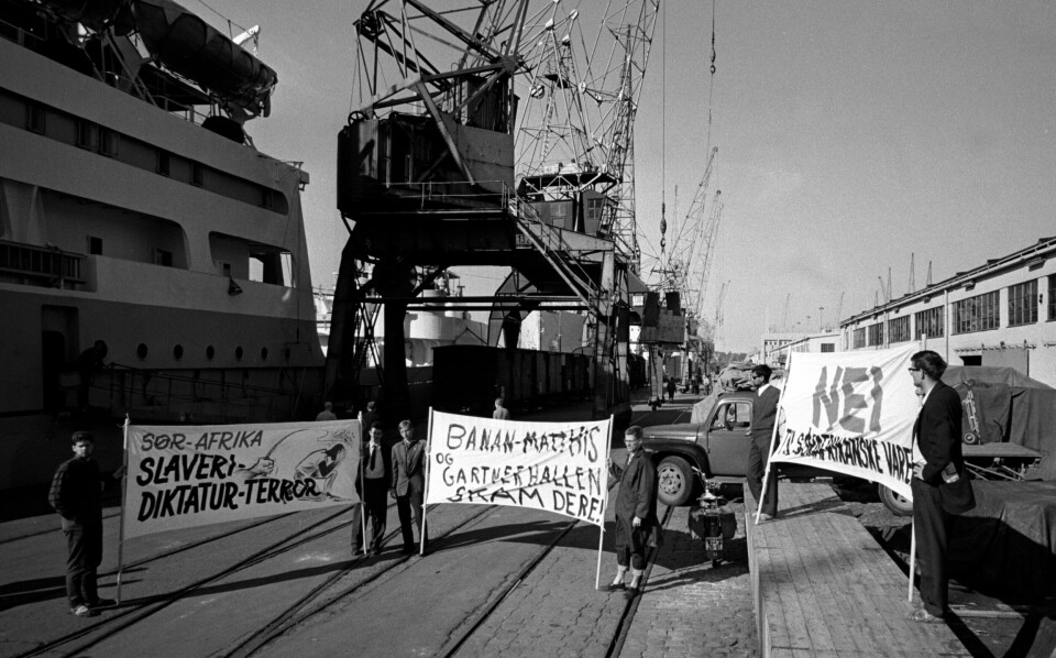 Demonstrasjon mot innføringen av sør-afrikanske varer på Oslo havn juni 1963. Plakatene viser «Sør-Afrika, Slaveri, Diktatur, Terror», «Banan-Mathiessen og Gartnerhallen skam dere» og «Nei til Sør-Afrikanske varer». Flere menn står samlet på havna med store plakater.