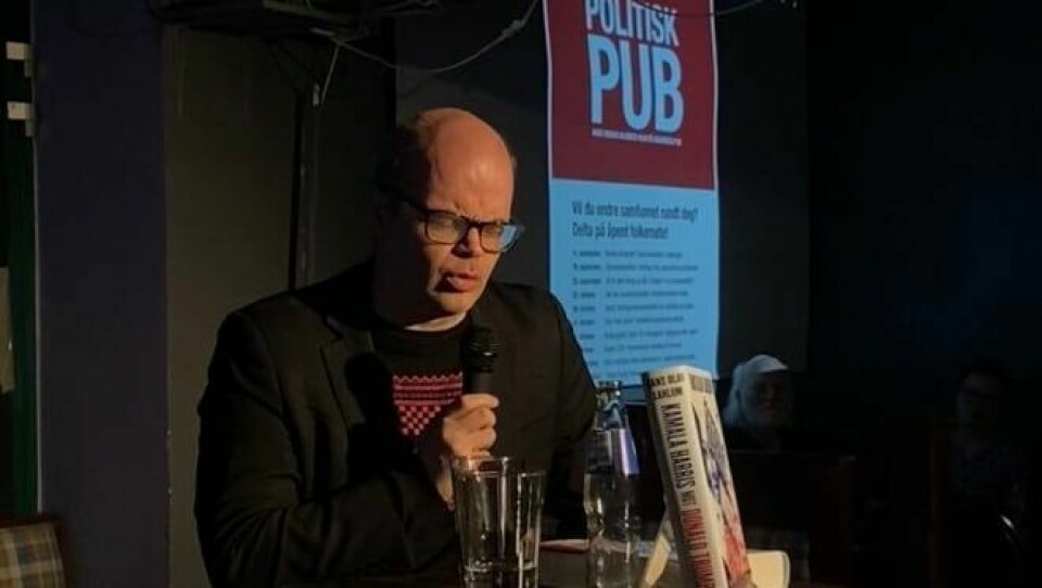 Hans Olav Lahlum er historiker og forfatter, sjakkformidler og samfunnsdebattant, og har fulgt amerikansk politikk i en årrekke. Dette er et mørkt bilde. Det er inne på en pub, og i en lysstråle sitter Hans Olav Lahlum, som har briller og lite hår. Han sitter ved et bord, har en mikrofon i hånden og bak ham er det skilt der det står 'Politisk Pub'