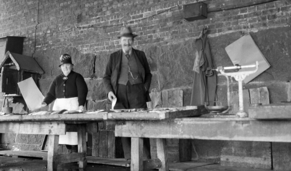 Fiskesalg ved Vaterlands bru i 1929, trolig på fiskebrygga. En mann og en dame stående bak en disk foran en murvegg.