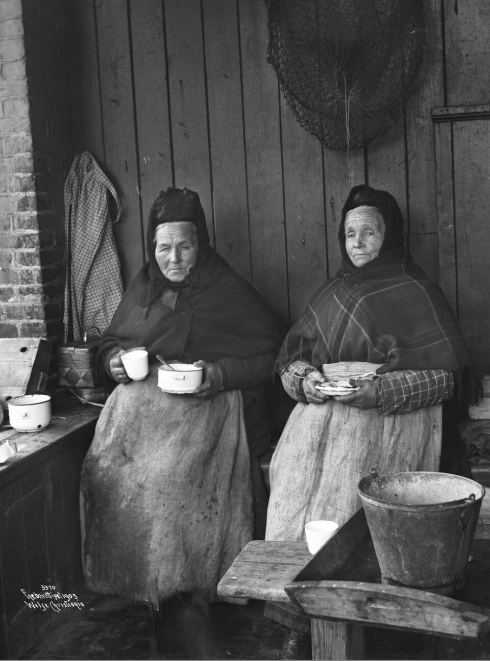 To fiskekoner tar lunsjpause i 1905. Disse to arbeida ved fiskebrygga i Bjørvika, ikke ved Vaterlandsbrua To fiskekoner sitter på en krakk og holder en bolle med mat i hånda.