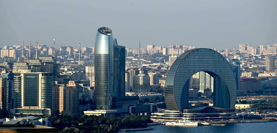 Dette fotografiet viser utsikten over den aserbajdsjanske hovedstaden Baku den 23. juli 2024. Flammer stiger opp fra en sandsteinsformasjon på en åsside på Absheron-halvøya nær Baku, Aserbajdsjans hovedstad, som forbereder seg på å være vert for klimakonferansen COP29 fra 11. til 22. november 2024. Det 'brennende fjellet' – Yanardag på aserbajdsjansk – næres av underjordisk gass som stiger til overflaten og antennes ved kontakt med oksygen. Baku er en grå by med høye bygg langs kysten. Forrest på bildet er det to veldig moderne bygg, den ene ser ut som en rakett og den andre er en svær halvsirkel.