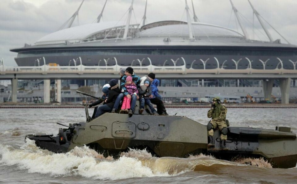 Folk leker seg under et militærshow mens den omstridte VM-arenaen i St. Petersburg reiser seg i bakgrunnen.