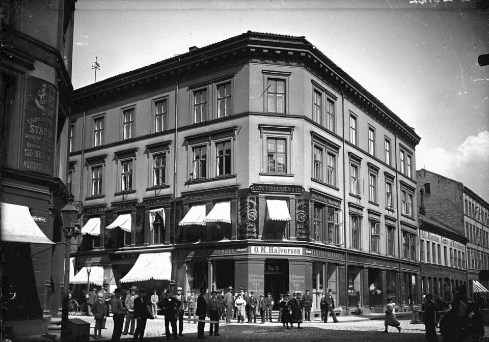Gatemiljøet i Skippergata på 1890-tallet. Bildet vise rSkippergata 27, 29 og 31, samt Karl Johans gate 5. Murbygningen består av fire etasjer. På hjørnet er det en inngang. På skiltet står det 'O. M Halvorsen brændevinshandel'.