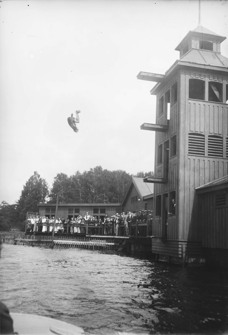 Svømmestevne 1916 på Filipstad. En deltaker hopper ut fra stupetårnet med hodet ned og knærne bøyd.