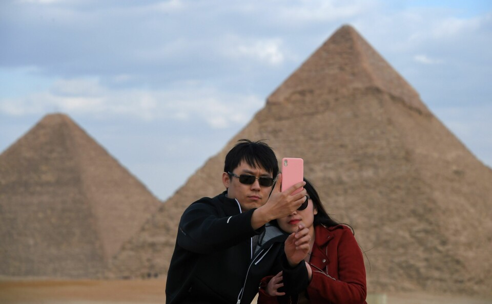 Turister med pyramidene til Khafre og Khufu i bakgrunnen. Et par poserer med mobiltelefonen foran to pyramider i Giza, Egypt. Himlen er blå og mannen har solbriller på.