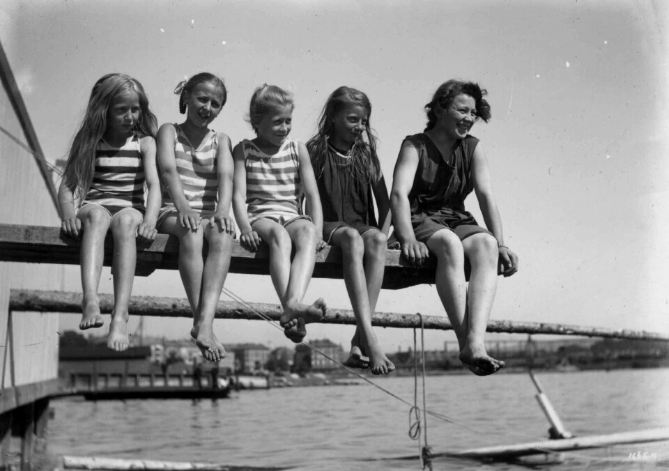 En kvinne med fire jenter sittende på en brygge Hasvold bad, ca. 1908. Tre av jentene har stripete badedrakt. Den siste jenta og kvinnen har mørke klær. Alle dingler med bena.