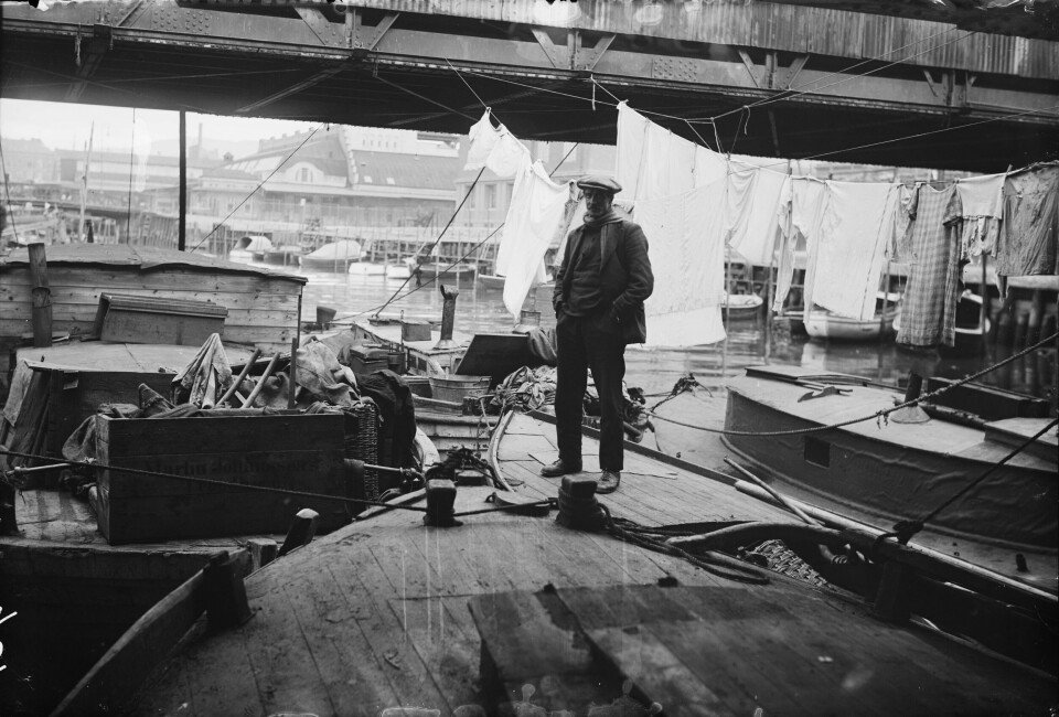 Klesvask og hverdagsliv på husbåter på Akerselva 1934. Flere båter samlet på vannet med klesvask i bakgrunnen og en mann i forgrunnen stående på en båt
