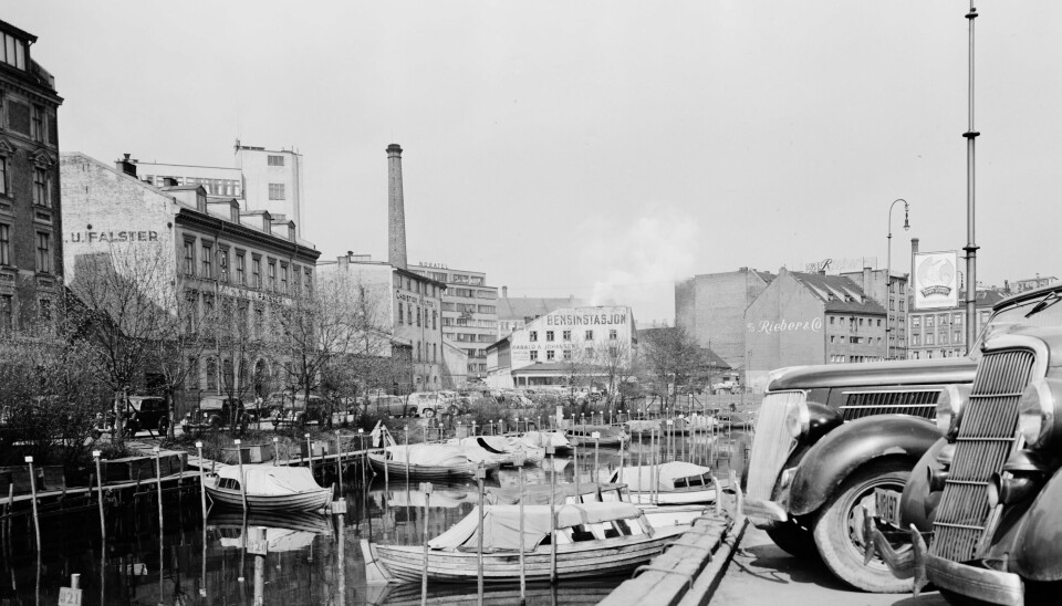 Akerselva sett fra Schweigaards bru mot Vaterlands bru i 1952. Båtliv og byliv. Biler stående langs kaia med bygninger i bakgrunnen.
