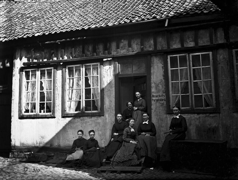 Kvinnen med korset om halsen er Lina Brøcher, flankert av sine ansatte. Kanskje er det pleiedattera hennes, Fredrikke som står ved siden av henne. Åtte personer står i trappa og foran inngangen til et hus.