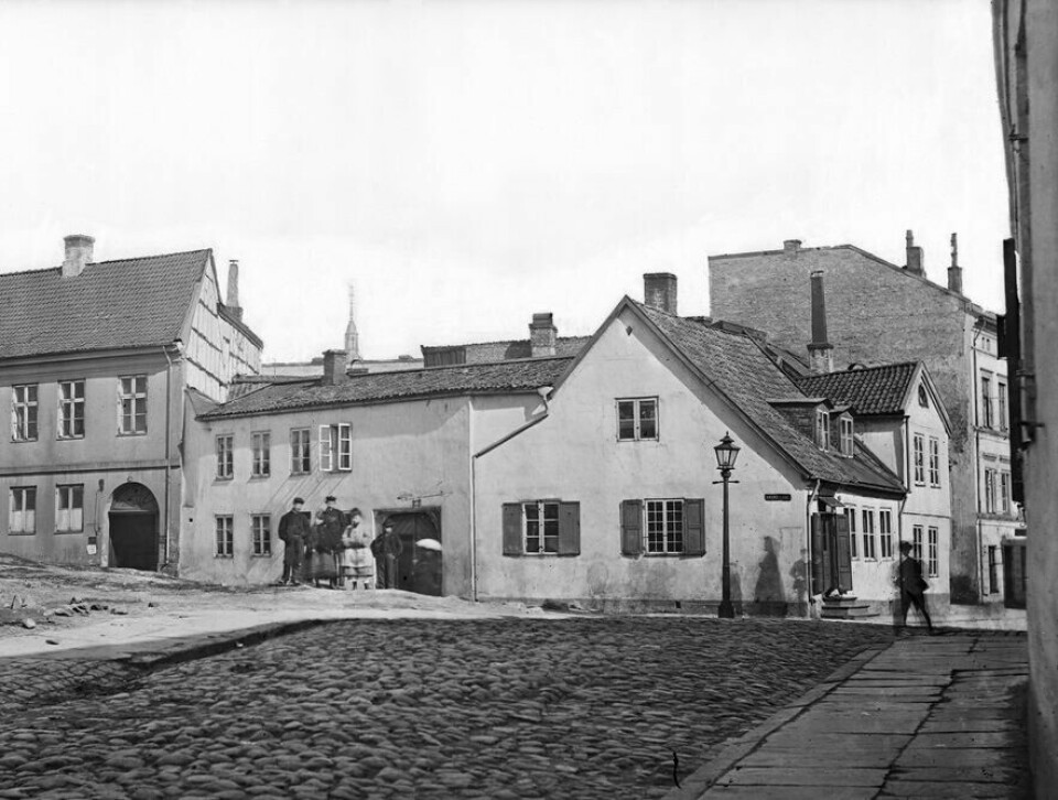 Her holdt fabrikken hus. Stortinget bygges til venstre. Foran en hvit murbygning står fire personer. En person med paraply skimtes foran døra.