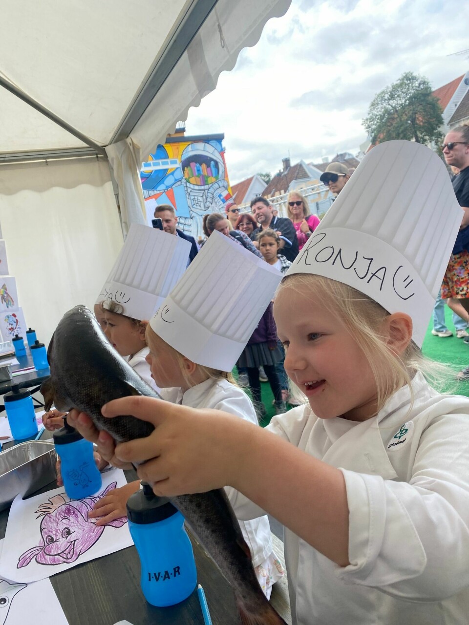 Ronja (5) er fornøyd med å få holde fisken. En jente på 5 år holder en fisk og har på en kokkehatt.