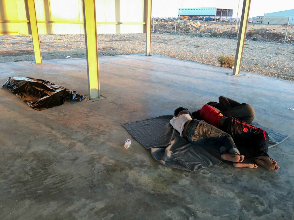 Overlevende hviler på et betonggulv på kysten av Libya fredag. I posen i bakgrunnen ligger ett av de rundt 150 menneskene som antas å ha druknet i Middelhavet.