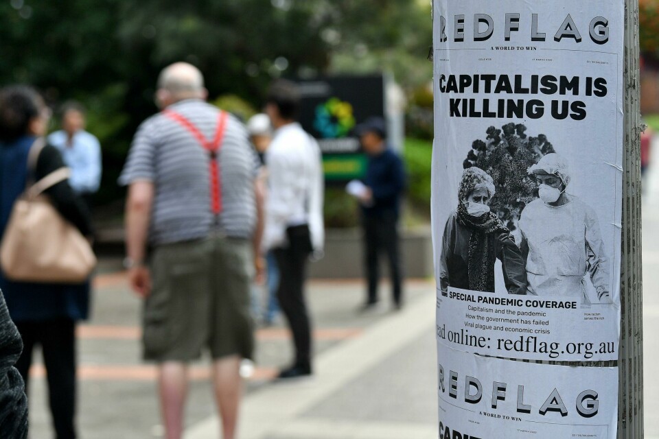 «Kapitalismen dreper oss», er budskapet på denne plakaten i Sydney, som ble satt opp etter at koronaen slo inn over Australia i mars og gjorde mange arbeidsløse der også. An anti-capitalist flyer is seen on a pole as people wait in a queue to receive benefit payouts, including unemployment and small business support as the novel coronavirus inflicts a toll on the economy, at a Centerlink payment centre in downtown Sydney on March 27, 2020. - The number of confirmed covid-19 cases in Australia passed the 3,000 mark on March 27, with the vast majority of infections in major east coast cities like Sydney, Brisbane and Melbourne. (Photo by Saeed KHAN / AFP)