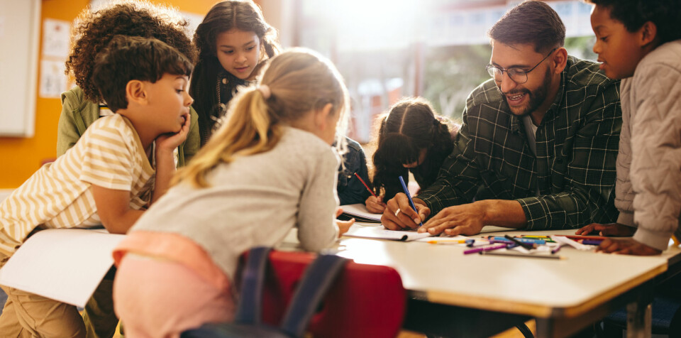 Tospråklig kompetanse blant de ansatte i lærerstaben og hos skoleassistenter og AKS-ansatte vil gjøre forståelse og tilhørighet til skolen lettere for mange elever, skriver Julie Remen Midtgarden. Illustrasjonsfoto. Group of elementary school kids stand around a table in a classroom, attentively following a lesson being taught by their teacher. Young students engaging in a fun activity of colouring and drawing.