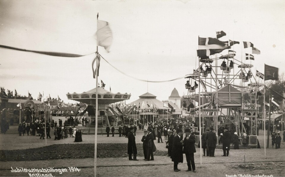 Bak flaggene sees Det russiske hjul ved Jubileumsutstillingen i 1914.(Ukjent/utstillingsforlaget/nb.no) Flere personer står blant en karusell og flere attraksjoner.